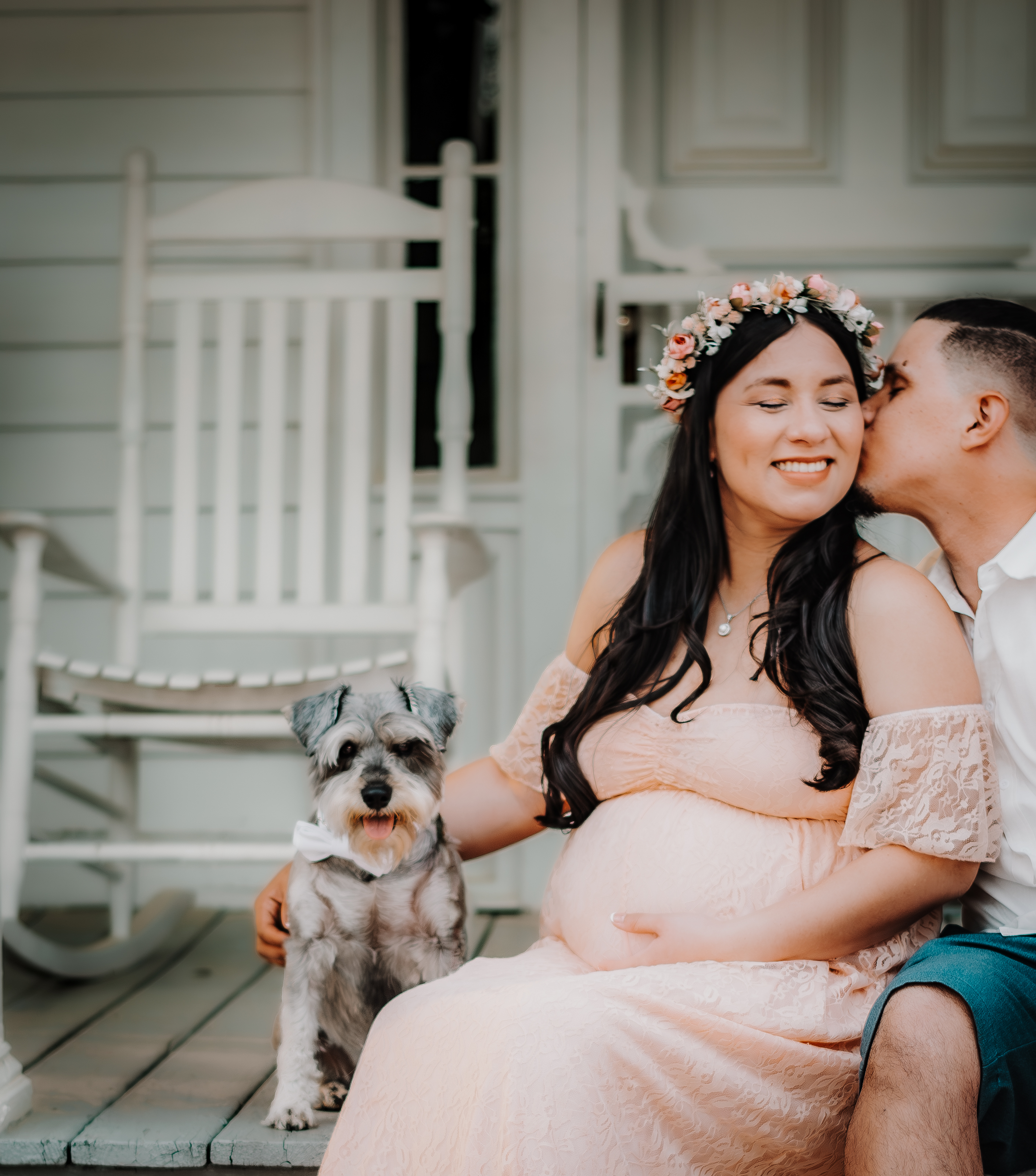 Man kisses his pregnant wife as they sit on the front porch of a house with their dog at McDaniel Farm Park.
