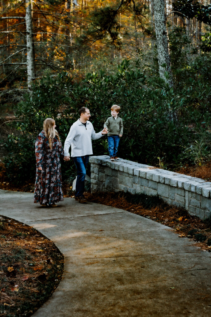 Mom and dad hold hands as little boy strolls along the wall, smiling back at them during a candid and movement based prompt of their fall family photo session. 