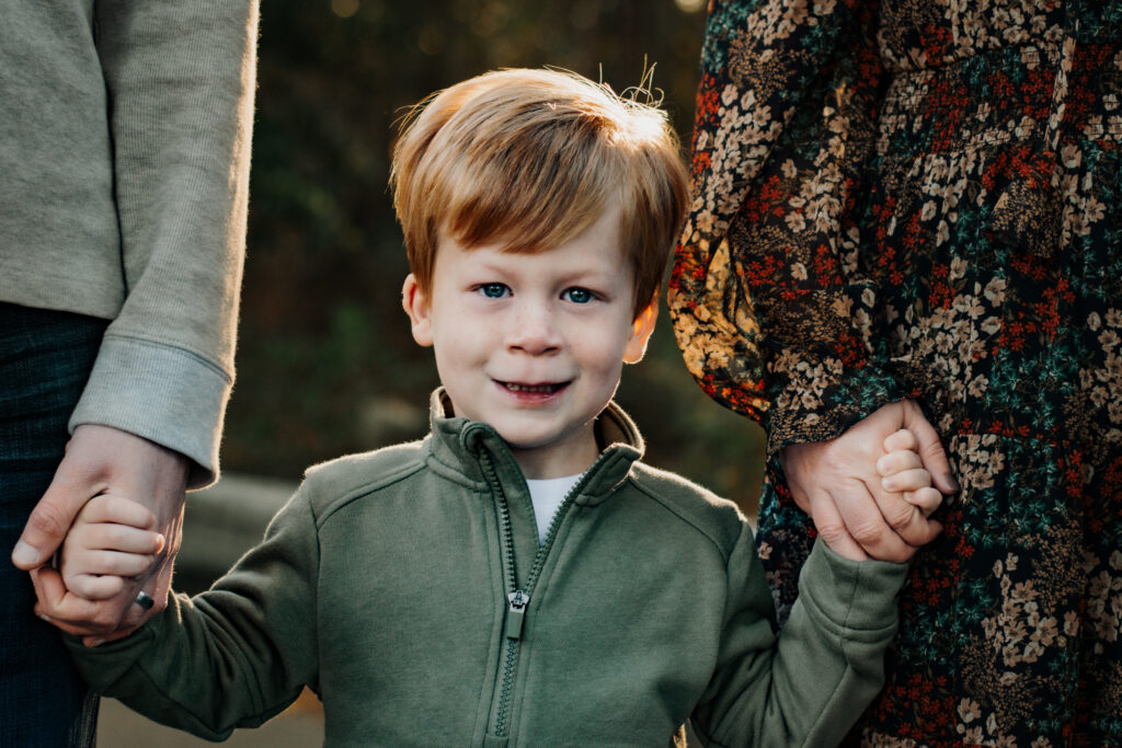 Little boy holds mom and dad's hands, eyes watery from the crisp fall air, smiling broadly into the camera during a family photo session in Loganville, Georgia. 