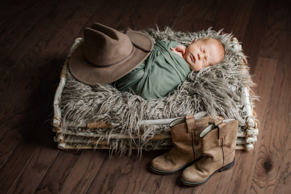 Newborn posed on gray fur in basket, squinting a bit with his dad's childhood cowboy hats and boots arranged around him.