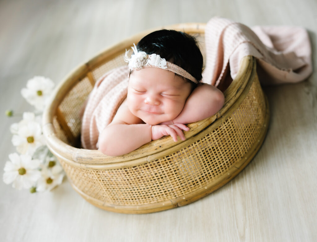 Smiling newborn poses chin on hands in a basket prop during an in-home newborn session.