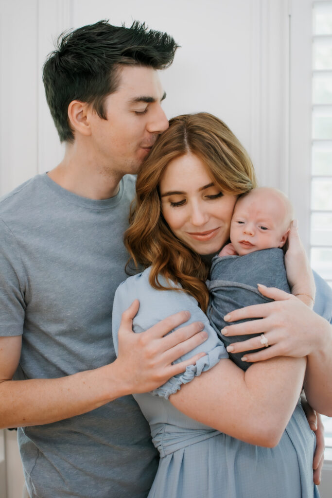 Dad kisses mom's head and she holds baby during in-home newborn session