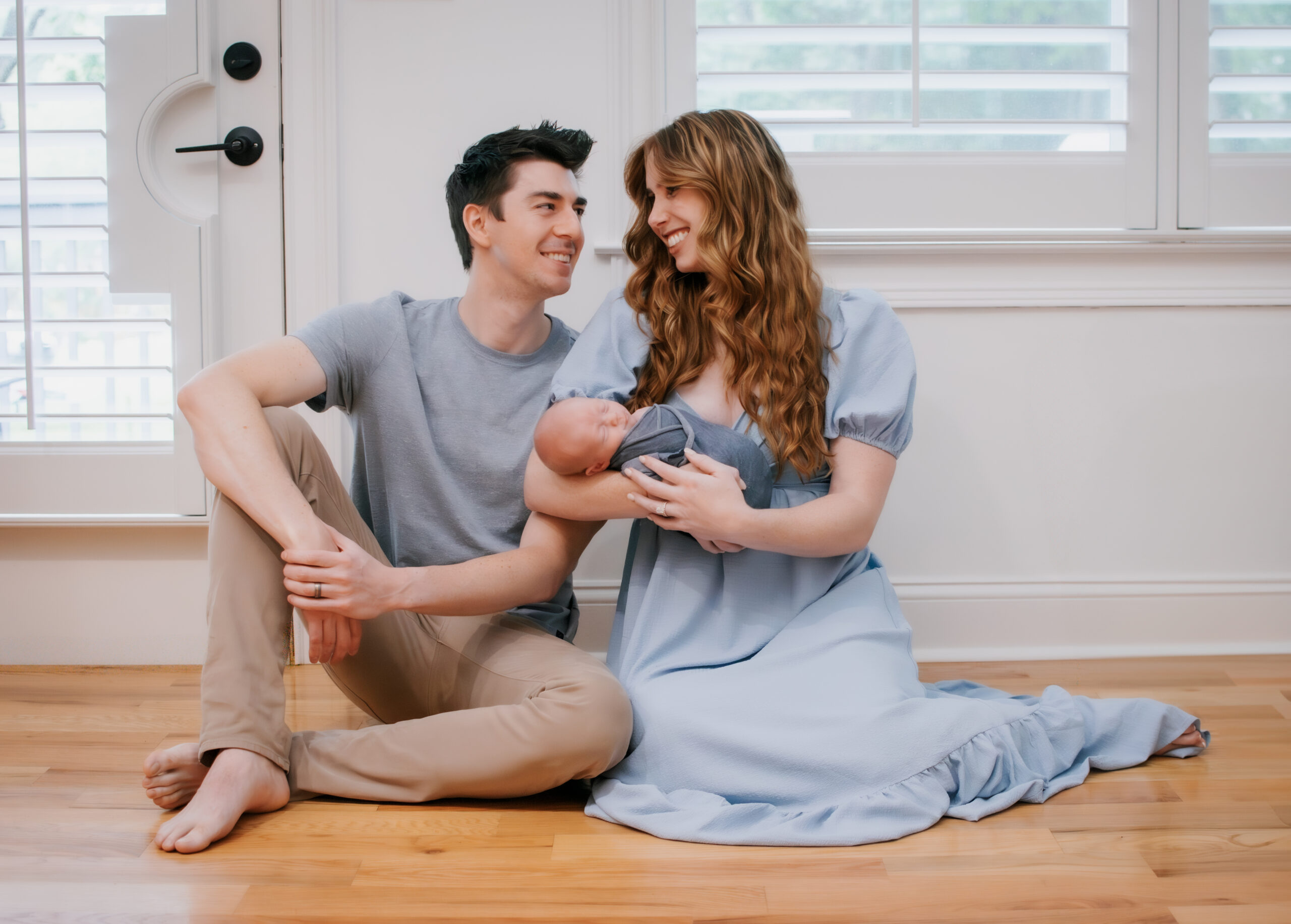 mom and dad sit on floor gazing at each other holding newborn during in-home session