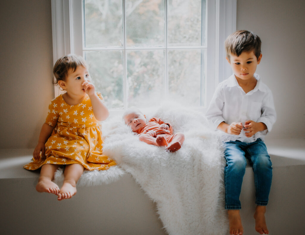 Brother and sister sit in widow with newborn sister during Atlanta photo session