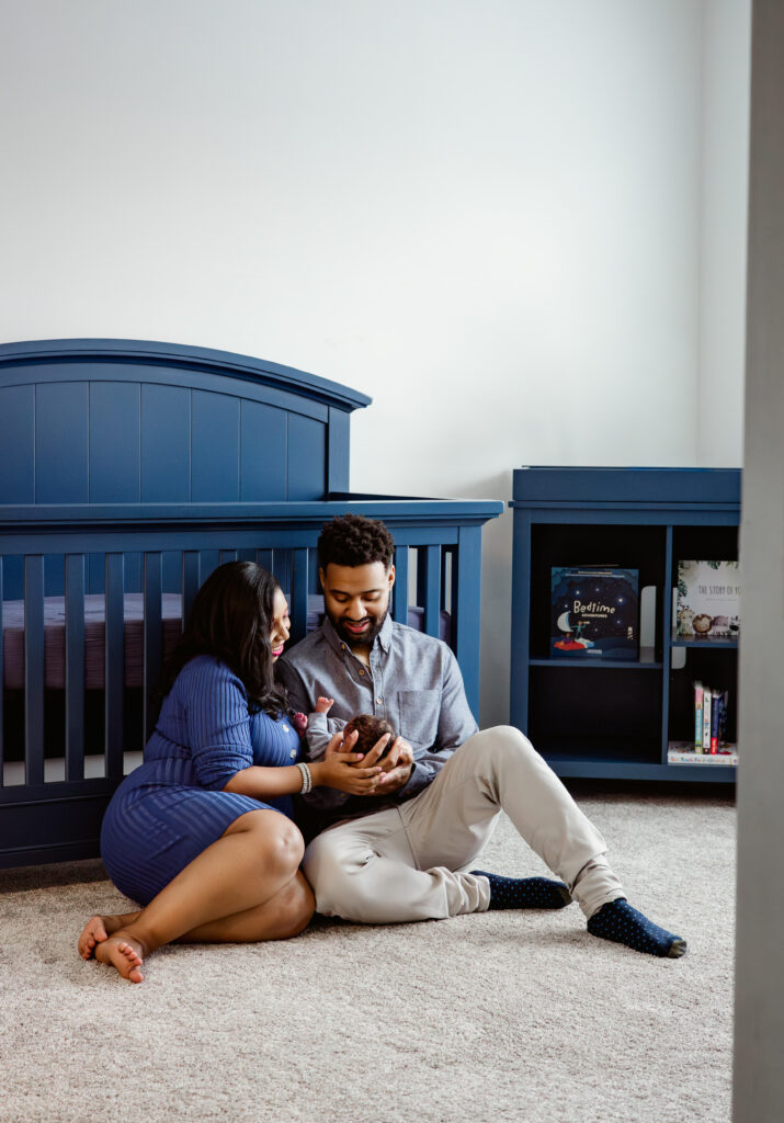 New parents sit in front of blue crib holding baby in newborn photo