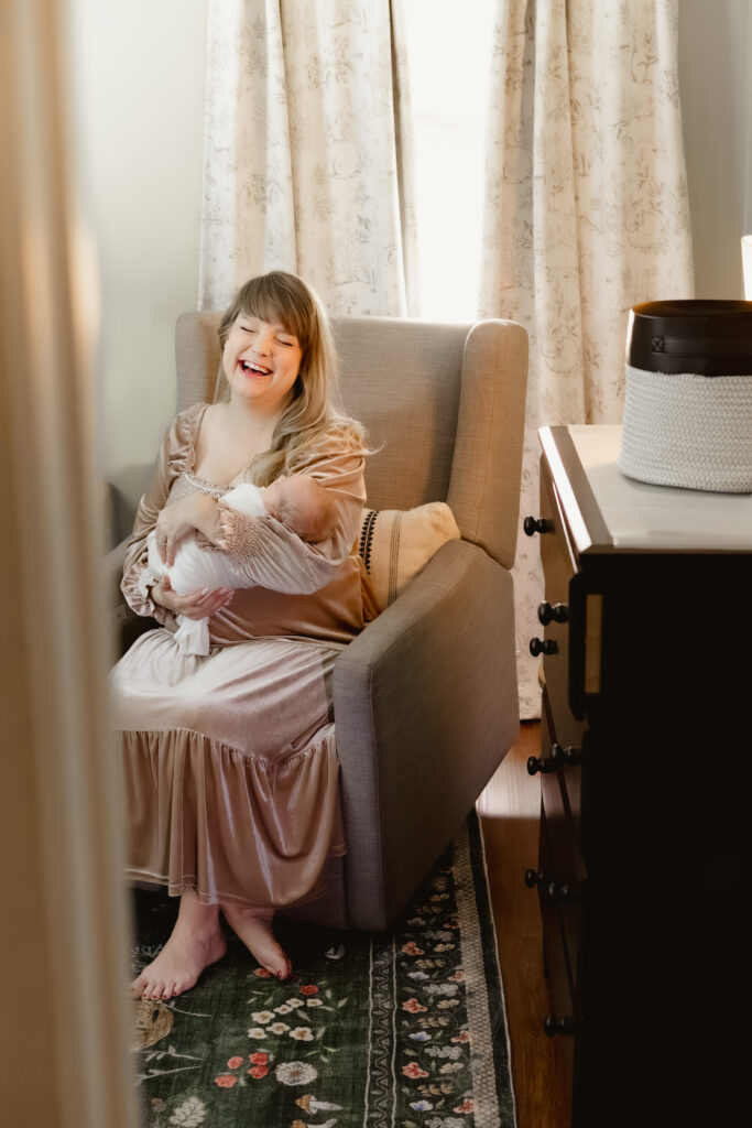Mom in rocking chair laughing while holding newborn baby.