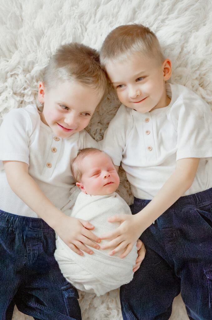 Twin brothers hold wrapped newborn on wool rug during in-home newborn session
