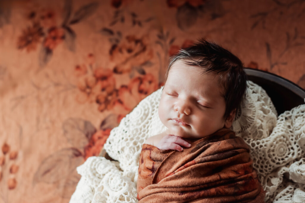 Newborn asleep in large bowl on lace, the background a rich rust floral - example of styling inventory for in-home newborn sessions in Atlanta. 