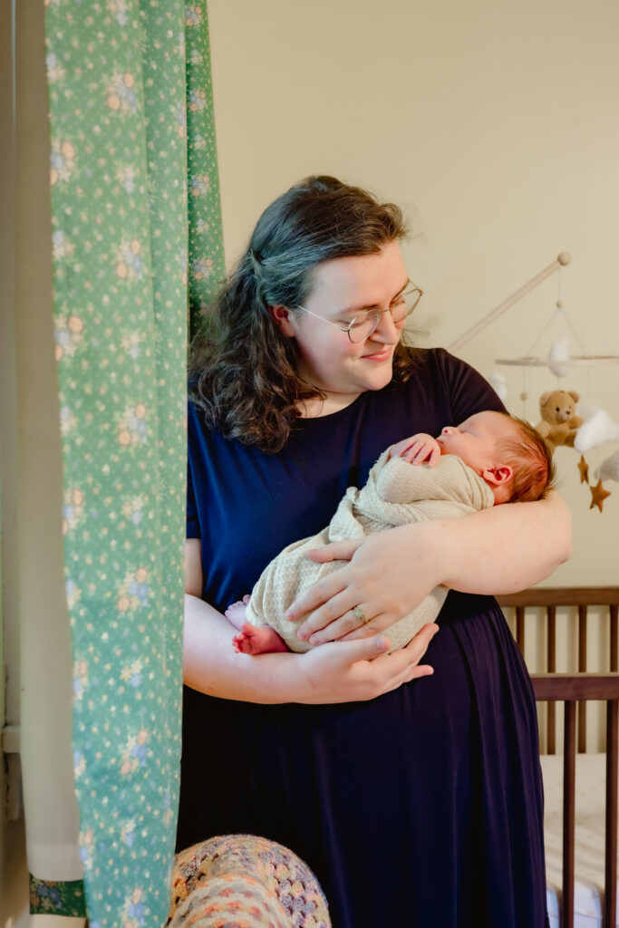 Mom holds her newborn son alongside the crib, the yellow nursery giving a warm hue to the image taken during a Tucker, Georgia newborn photo session. 