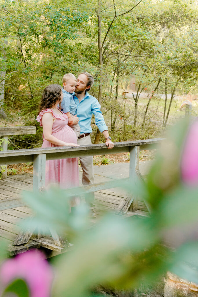 Expecting mama, dad and toddler pose candidly on a bridge for maternity session at golden hour