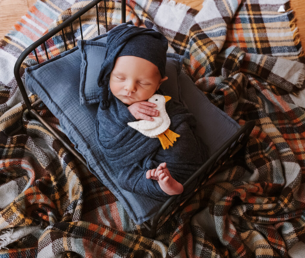 Newborn baby boy sleeps in blue with a cap and wrap on a small bed atop a floral blanket during an in-home newborn photography session in Lilburn, Georgia.