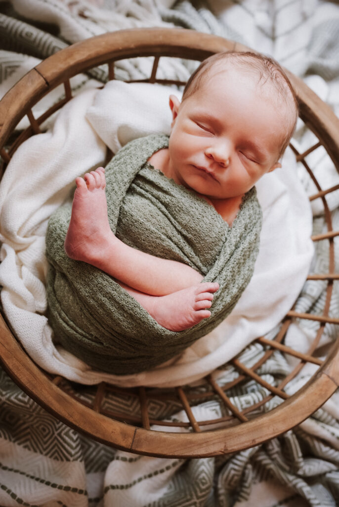 Little boy sleeps curled in a green wrap, toes peeking, resting in a small basket during an Atlanta in-home newborn session.