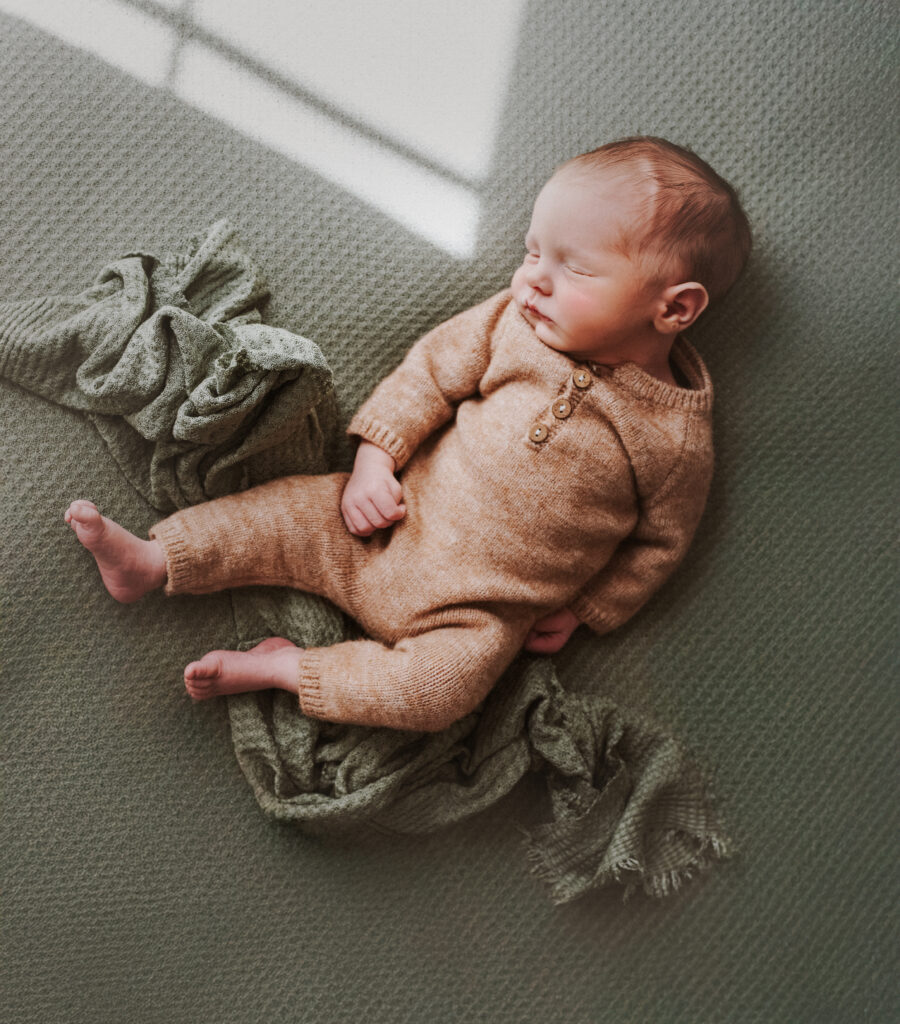 Unposed Newborn sleeps peacefully on a green backdrop, wearing a tan romper during his Atlanta Georgia newborn session in the family home.